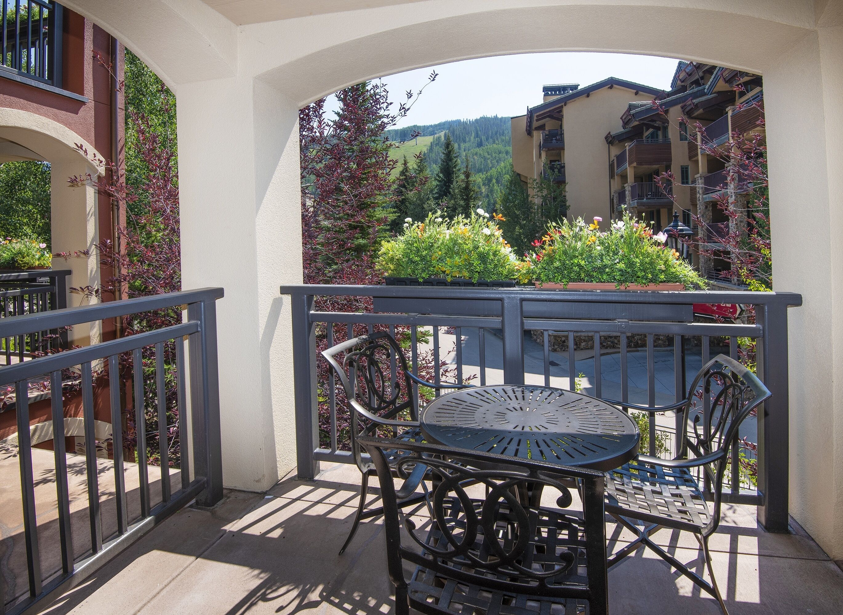A balcony with a table and chairs, surrounded by potted plants and a view of buildings and hills.