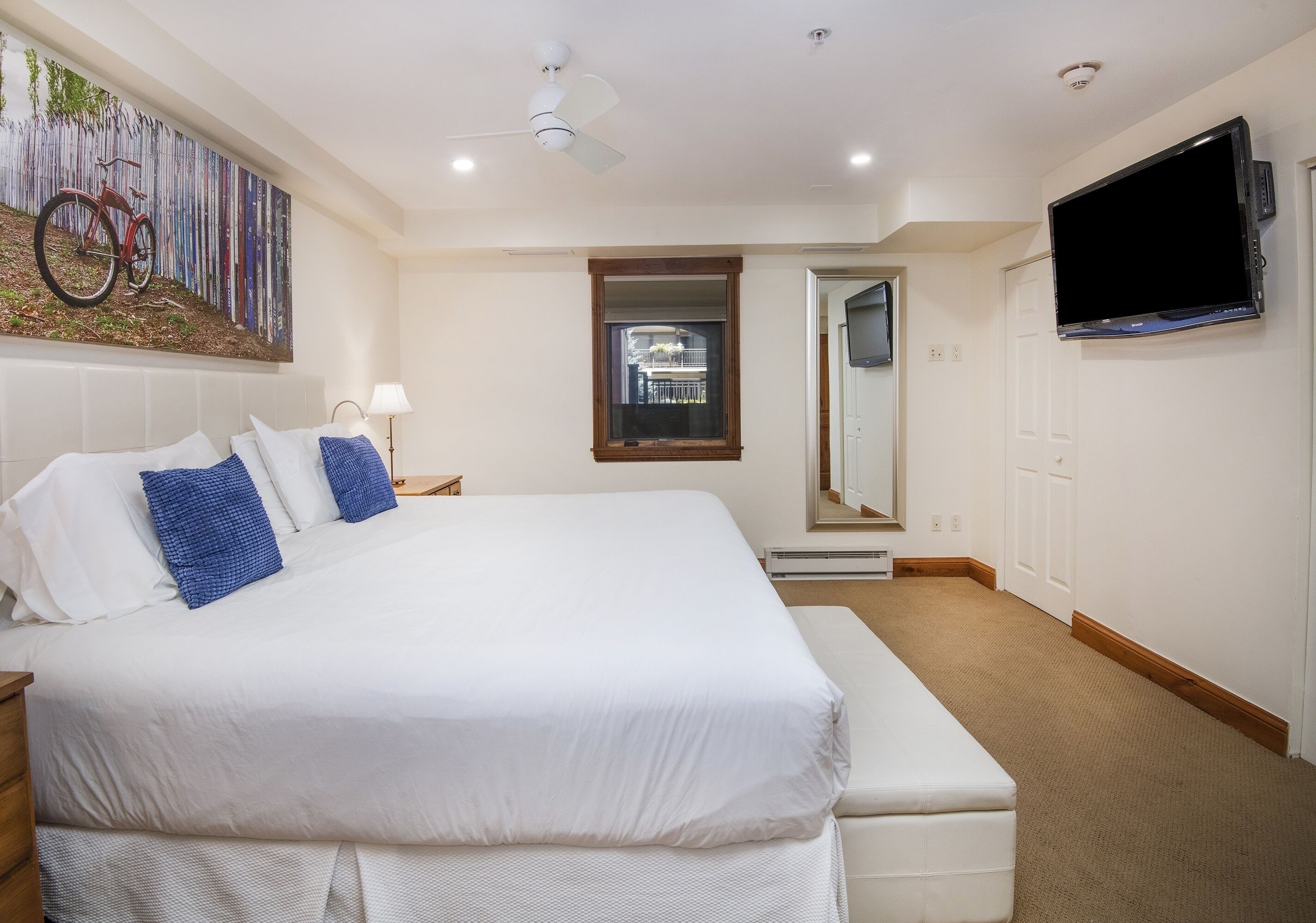 A hotel room with a large bed, a flat-screen TV mounted on the wall, a ceiling fan, and a window with a view of a building.