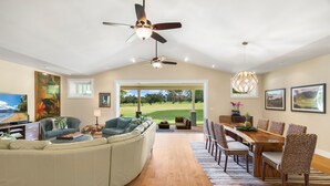 A spacious living room with a curved sofa, a dining area with a wooden table and wicker chairs, and a large window offering a view of a golf course. - Hale Alaula, Elegant Hawaiian Home at exclusive Sunset Drive (Princeville)