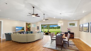 A spacious living room with a large L-shaped sectional sofa, a dining area with a wooden table and chairs, and a view of a golf course through the glass door. - Hale Alaula, Elegant Hawaiian Home at exclusive Sunset Drive (Princeville)