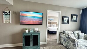 Un salon avec une grande télévision diffusant une scène de plage, un meuble turquoise, un canapé à motifs et un lit donnant sur la pièce.