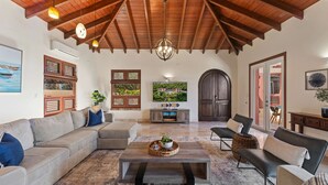 A spacious living area with a high wooden ceiling, a large sectional sofa, a coffee table with decorative items, and a television mounted on the wall. - Private Villa*MagensBay *Ocean front* Private Pool (St. Thomas)