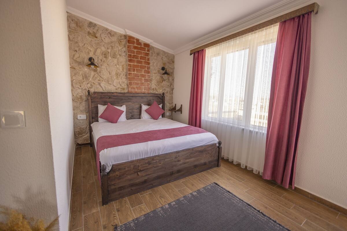 Ein Schlafzimmer mit Steinwand, einem hölzernen Bett, roten Vorhängen und Blick ins Freie.