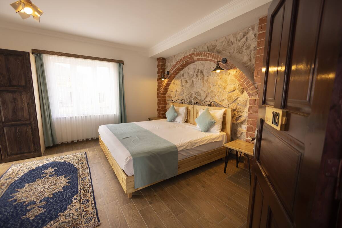 Ein Schlafzimmer mit Kamin aus Stein, einem Bett mit Kissen, einem Nachttisch aus Holz und einem großen Fenster mit Vorhängen.