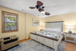 A bedroom with a bed, a ceiling fan, a window with blinds, a bedside table with a lamp, and a suitcase.