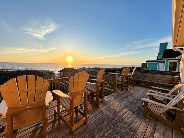 A wooden deck with Adirondack chairs facing a sunset over the ocean.