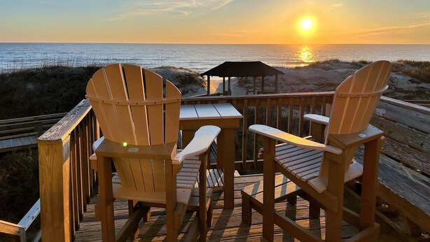 Eine Holzterrasse mit zwei Adirondack-Stühlen und einem Tisch, mit Blick auf einen Strand bei Sonnenuntergang.
