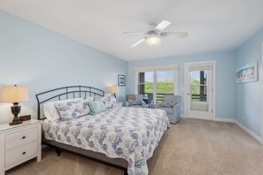 A bedroom with a bed, two chairs, a nightstand, a ceiling fan, and a window with a view.