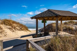 A wooden beach shelter with a sloped roof, situated on a sandy beach with dunes and the ocean in the background.