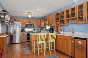 A modern kitchen with wooden cabinets, stainless steel appliances, and a central island with chairs. - Tidy Duck-Semi-oceanfront with views, private pool, hot tub, steps to the beach! (Duck)