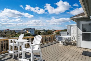A wooden deck with white chairs and a table, overlooking a residential area with houses and a clear sky. - Tidy Duck-Semi-oceanfront with views, private pool, hot tub, steps to the beach! (Duck)
