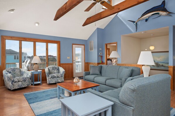 A spacious living room with a large gray sofa, two patterned armchairs, a wooden coffee table, and a view of the outside through large windows.