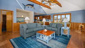 A spacious living room with a large gray sectional sofa, a wooden coffee table, and a ceiling fan. A kitchen area is visible in the background with wooden cabinets and a large window. - Tidy Duck-Semi-oceanfront with views, private pool, hot tub, steps to the beach! (Duck)