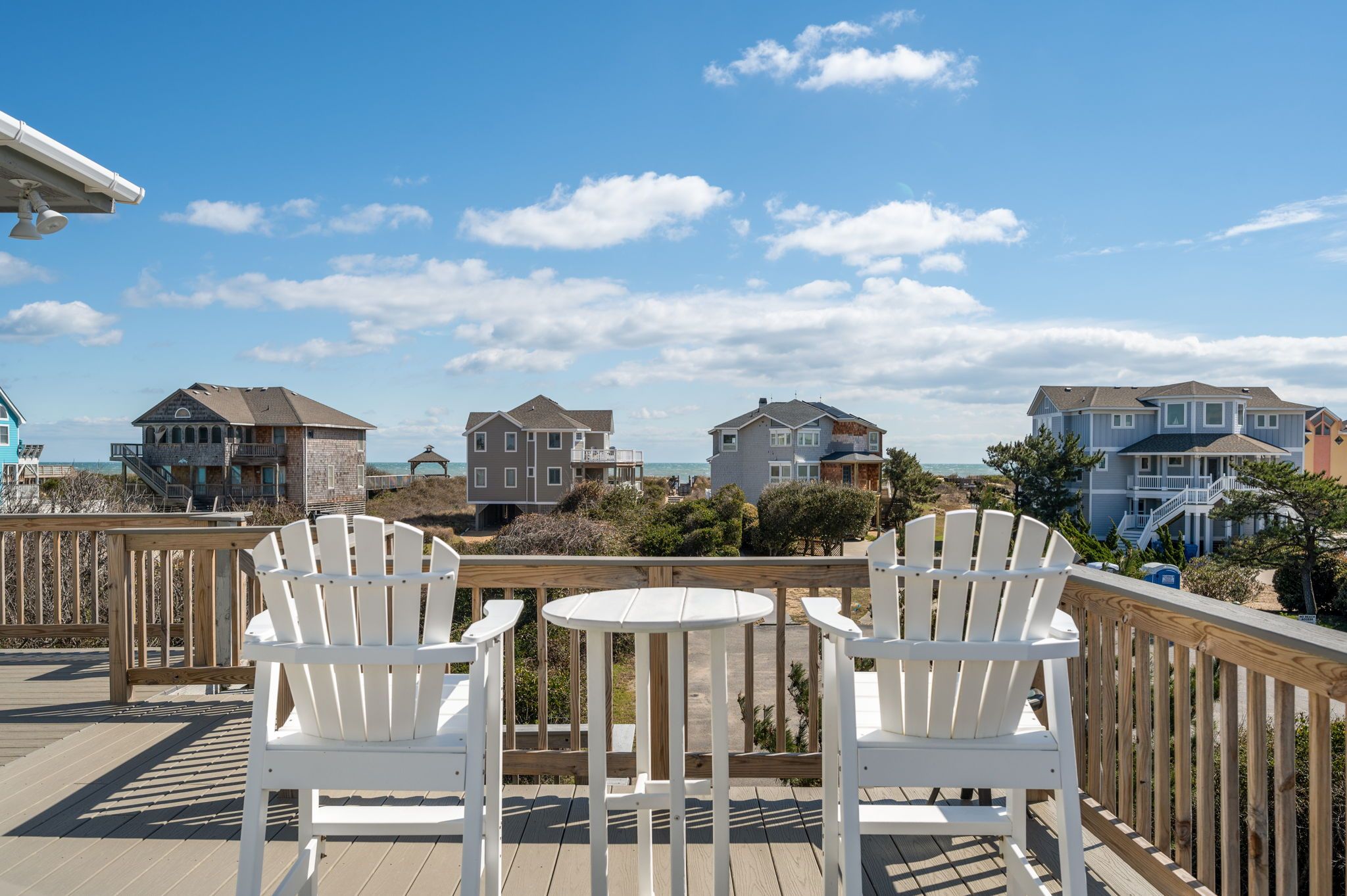 A wooden deck with two white chairs and a small round table, overlooking a beachfront with several houses.
