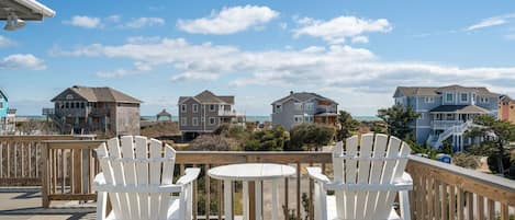 A wooden deck with two white chairs and a small round table, overlooking a beachfront with several houses.