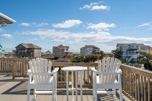 Une terrasse en bois avec deux chaises blanches et une petite table ronde, donnant sur une plage bordée de plusieurs maisons.