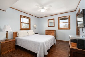 Un dormitorio con una cama, mesitas de noche de madera, un escritorio con una computadora, un ventilador de techo y dos ventanas con marcos de madera.