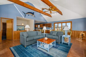 A spacious living room with a large gray sectional sofa, a wooden coffee table, and a ceiling fan. A kitchen area is visible in the background with wooden cabinets and a large window.