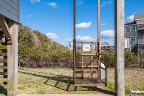 A wooden swing chair with a sign, surrounded by a grassy area and a wooden fence, with houses in the background. - Tidy Duck-Semi-oceanfront with views, private pool, hot tub, steps to the beach! (Duck)