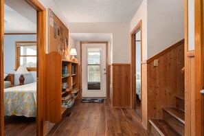 A hallway with wooden flooring, a door leading to a room with a bed and a lamp, and a shelf with books and a lamp. - Tidy Duck-Semi-oceanfront with views, private pool, hot tub, steps to the beach! (Duck)
