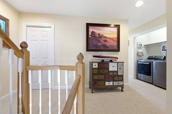 A hallway with a wooden staircase, a white door, a wooden cabinet with drawers, a painting, and a kitchen area with appliances.