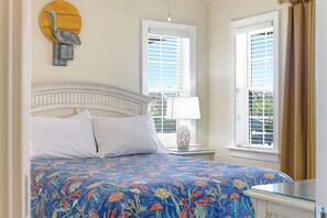 A bedroom with a bed, two windows with blinds, a nightstand with a lamp, and a wall decoration.