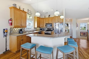 A kitchen with wooden cabinets, a central island with bar stools, a microwave, a refrigerator, and a coffee maker. - Queen Anne's Retreat: Oceanside, ocean views, private pool, hot tub & rec room (Nags Head)