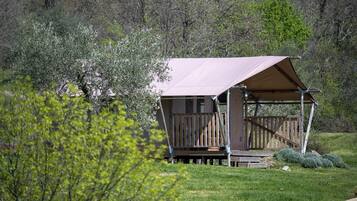 A small, single-story cabin with a wooden deck and a covered porch, surrounded by greenery and trees.
