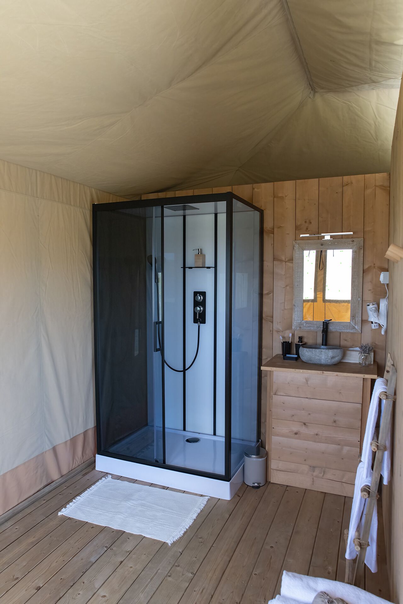 Une chambre moderne, aménagée comme une tente, avec une douche en verre, une commode en bois avec un lavabo et un miroir.
