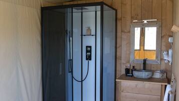 Une chambre moderne, aménagée comme une tente, avec une douche en verre, une commode en bois avec un lavabo et un miroir.