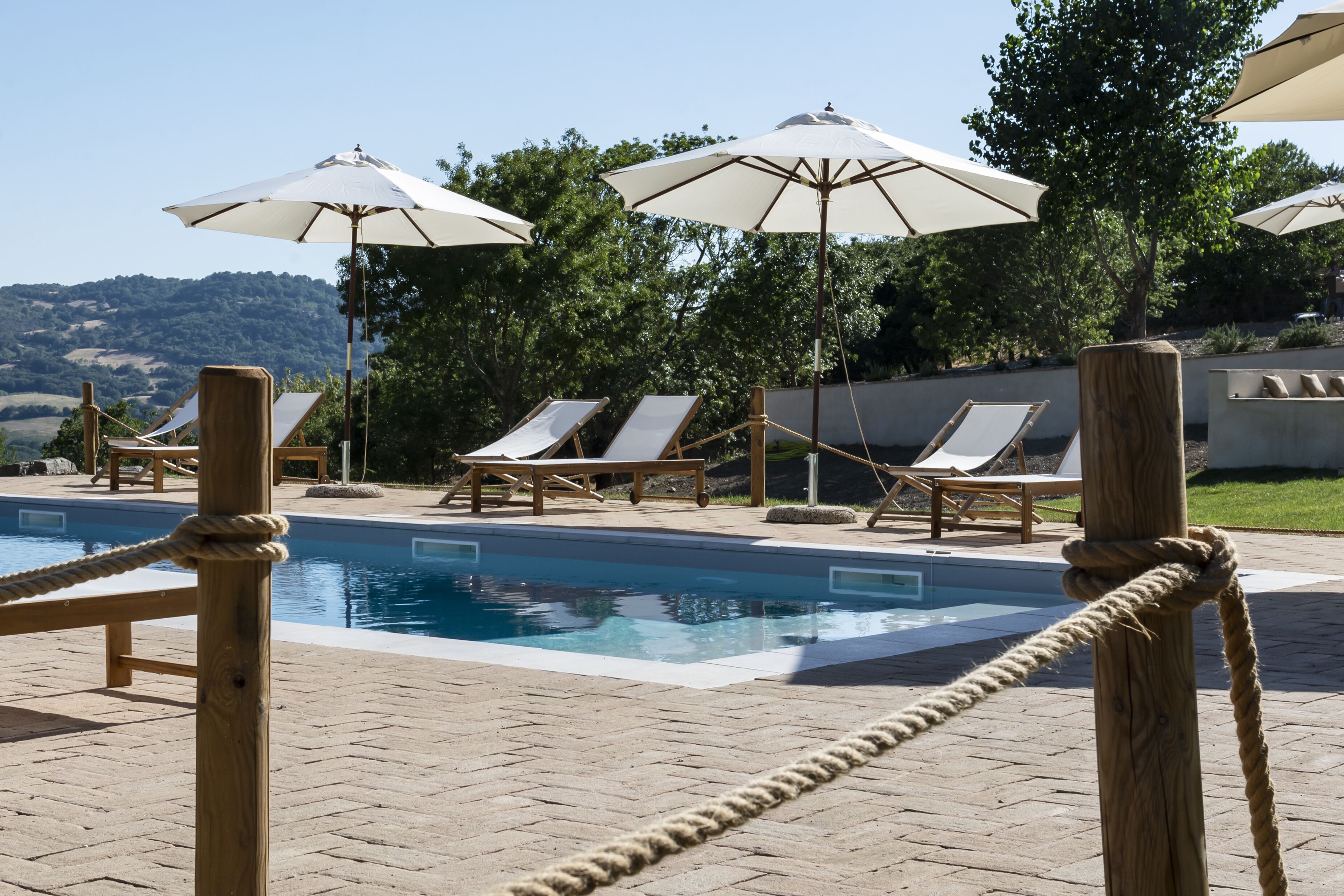 Ein Poolbereich mit Liegestühlen und Sonnenschirmen, umgeben von Bäumen und Blick auf Hügel.