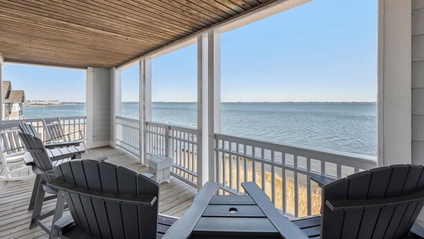 A covered deck with Adirondack chairs and a table, overlooking a beach and the ocean. - Whispering Sound: Waterfront | Pool/Gym Access (Manteo)