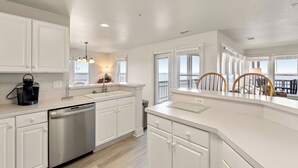A modern kitchen with white cabinetry, a dishwasher, a coffee maker, and a view of the ocean through large windows. - Whispering Sound: Waterfront | Pool/Gym Access (Manteo)