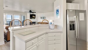 A modern kitchen with white cabinetry, a stainless steel refrigerator, and a built-in oven. There are wooden chairs and a dining table in the adjacent dining area. - Whispering Sound: Waterfront | Pool/Gym Access (Manteo)