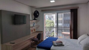 Una habitación de hotel con una cama, un televisor montado en la pared, un escritorio con una silla y un balcón con vistas a edificios y zonas verdes.