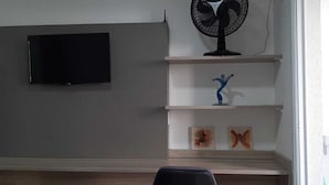 Una habitación con televisión de pantalla plana en la pared, un ventilador de pie y una estantería con adornos.