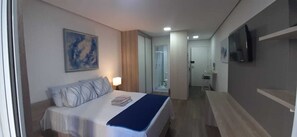 Une chambre d’hôtel avec un lit, une lampe de chevet, un téléviseur fixé au mur et une salle de bain visible à travers une porte ouverte.