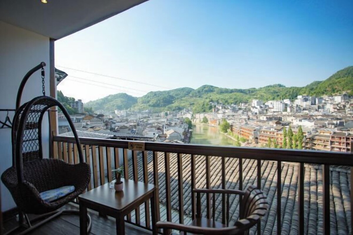 A balcony with a wicker chair and table, overlooking a cityscape with buildings and greenery.