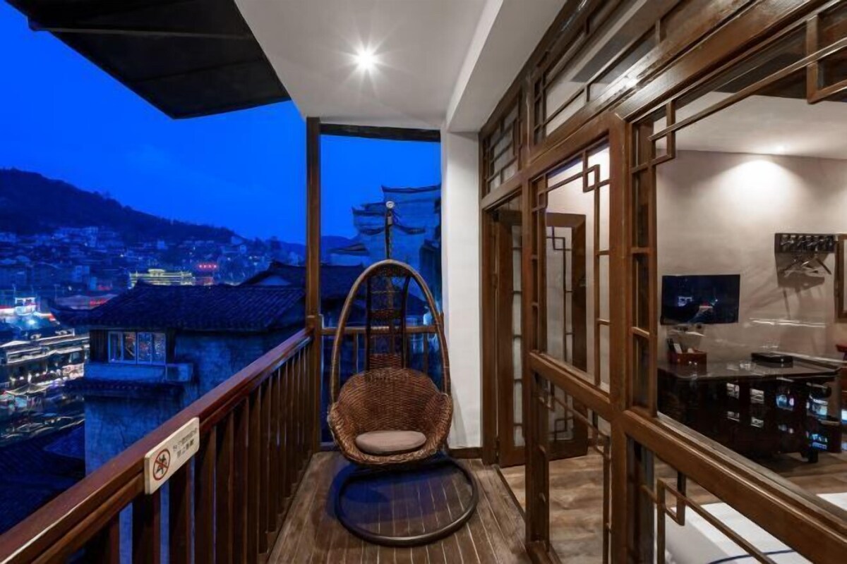 A balcony with a wicker chair, a TV, and a view of a cityscape at dusk.