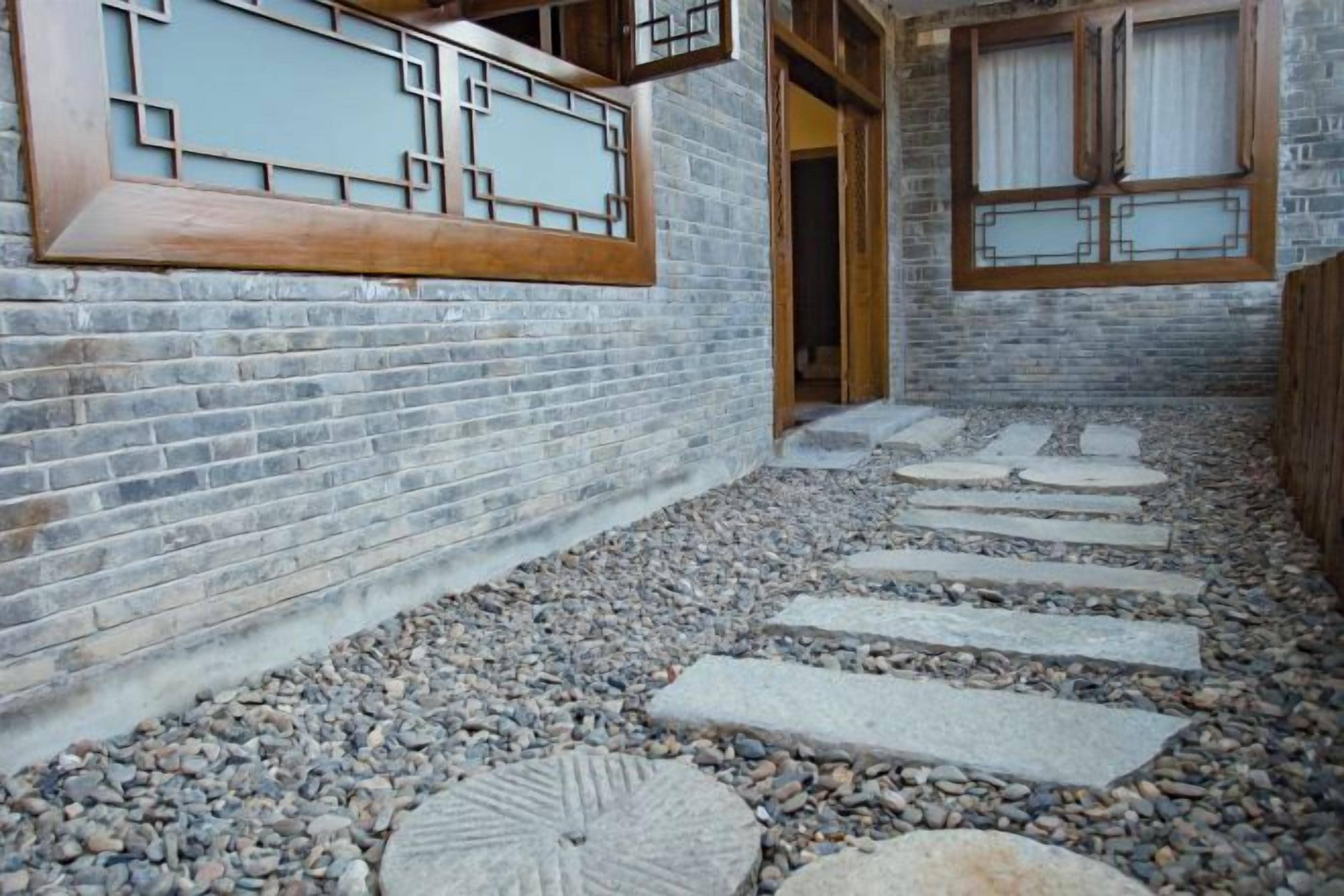A courtyard with a stone pathway leading to a wooden door, surrounded by brick walls and a traditional window design.