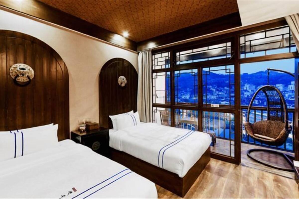 Une chambre d’hôtel avec deux lits, une grande fenêtre donnant sur un beau paysage, un plafond en bois et un fauteuil suspendu près de la fenêtre.