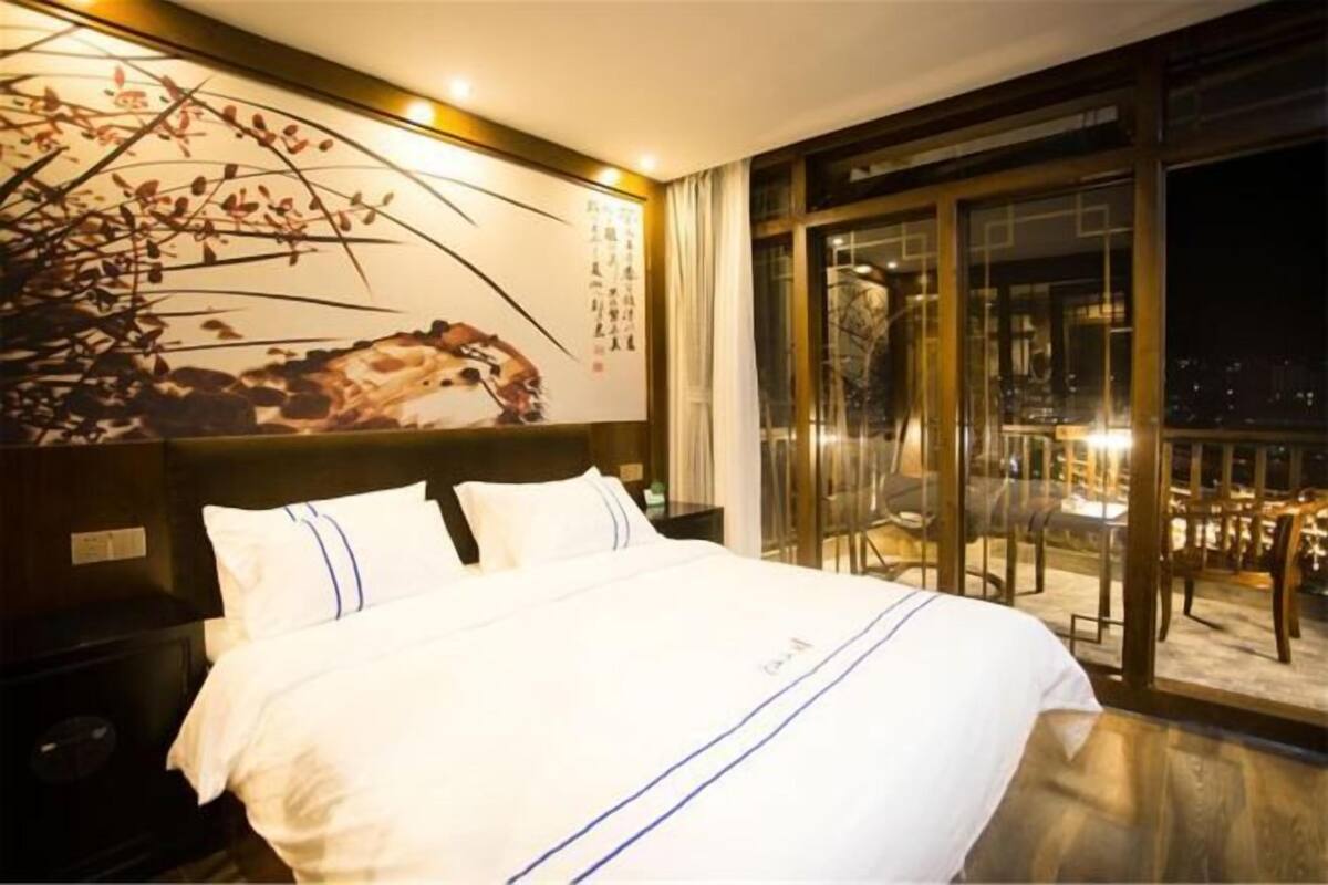 Une chambre d’hôtel avec un grand lit, une table de chevet, une œuvre d’art accrochée au mur et une vue sur la ville grâce à de grandes fenêtres.