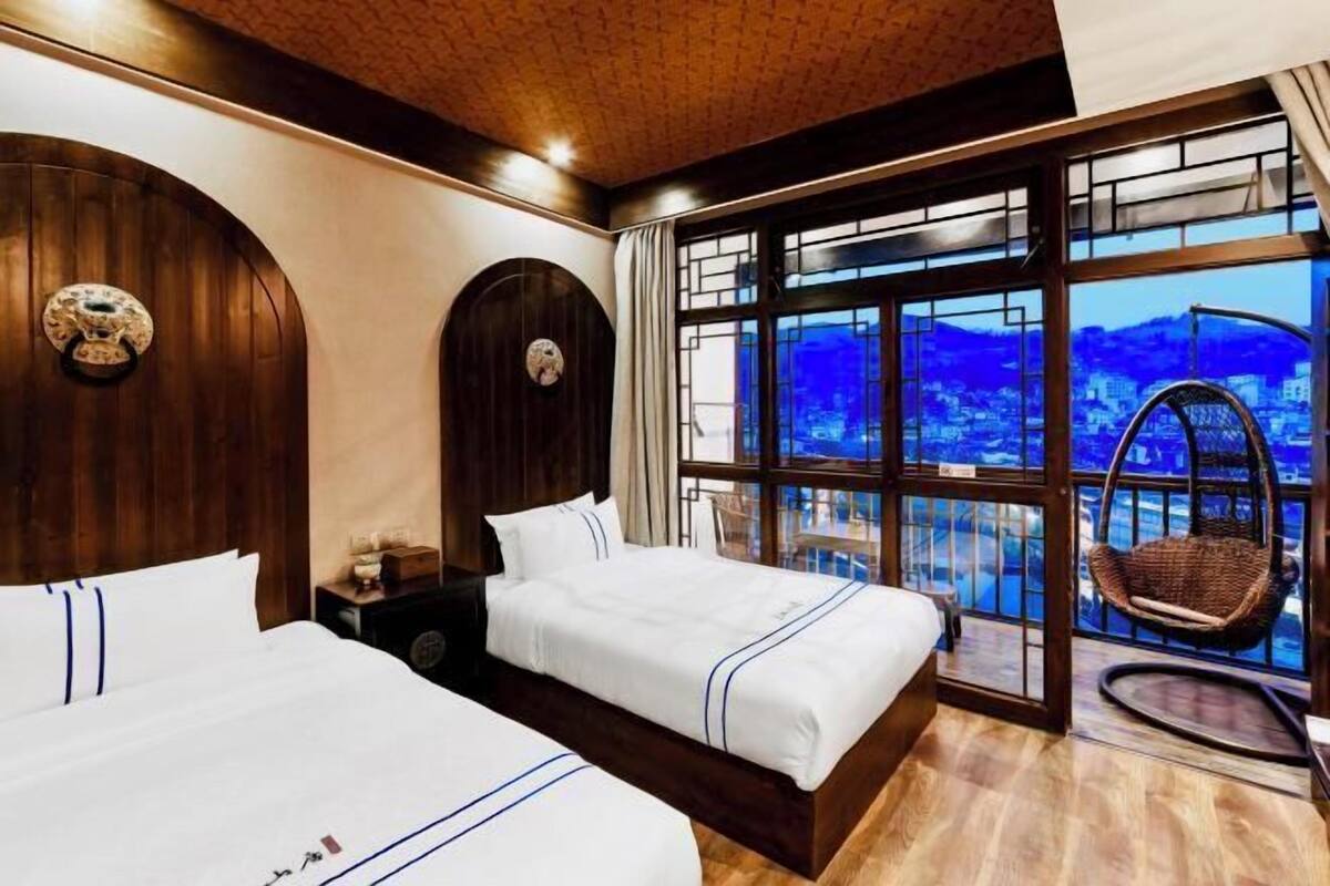 Une chambre d’hôtel avec deux lits, une grande fenêtre donnant sur un paysage urbain et un fauteuil en osier près de la fenêtre.