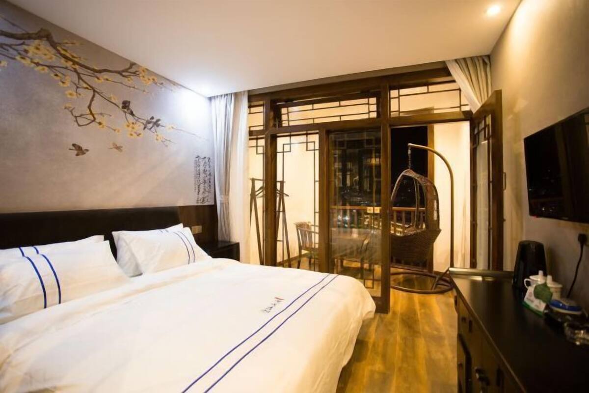 Une chambre d’hôtel avec un grand lit, une fresque murale, une porte-fenêtre coulissante donnant sur un balcon et un téléviseur posé sur la commode.