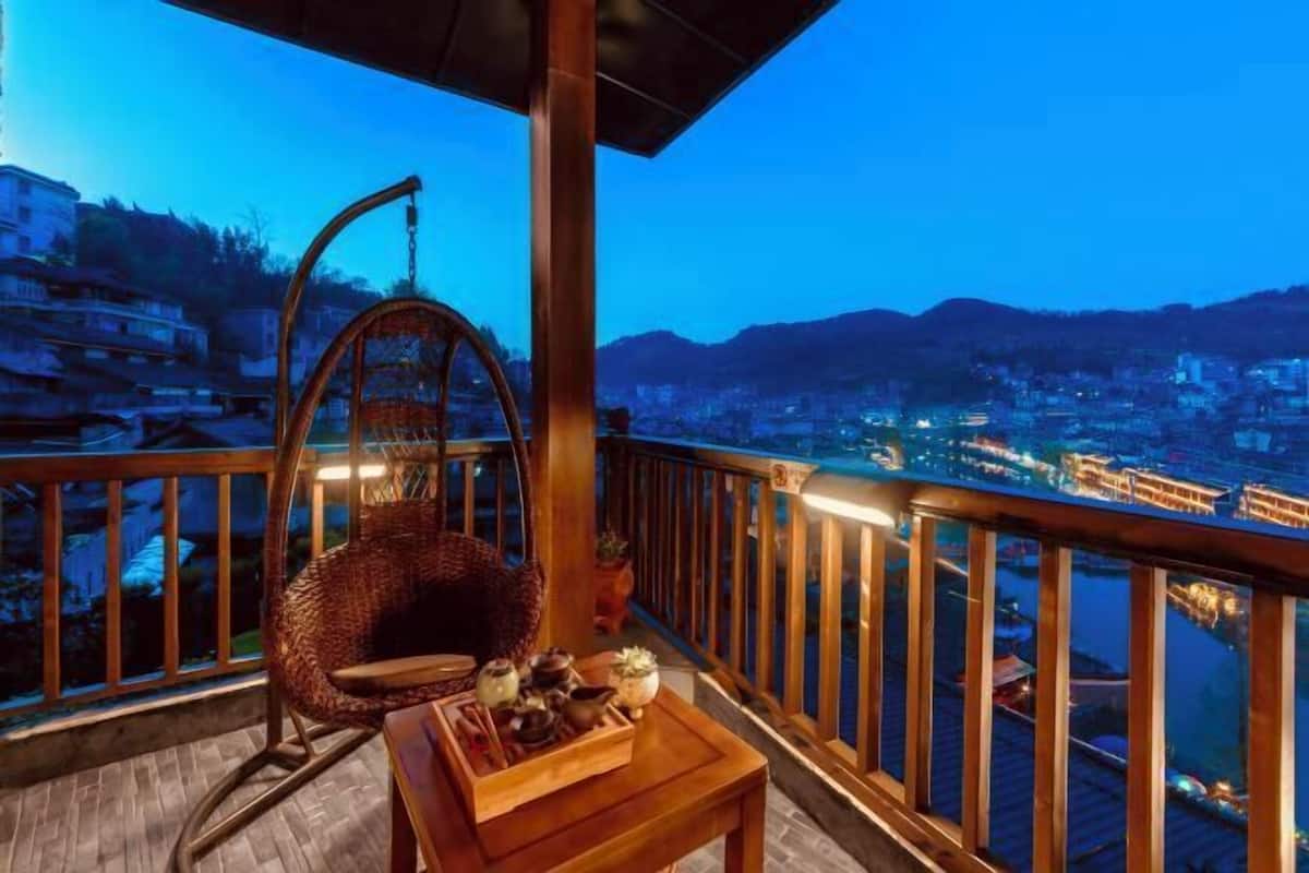 Un balcon avec un fauteuil en osier, une petite table agrémentée d’objets décoratifs et une vue sur la ville au crépuscule.