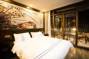 A hotel room with a bed, a nightstand, a wall with artwork, and a balcony with chairs. - Wangjiang Shanju Guesthouse (Fenghuang County)
