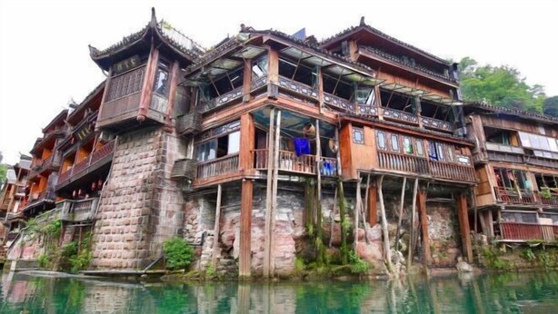 A traditional multi-story building with wooden balconies and a stone base, situated by a body of water. - Wangjiang Shanju Guesthouse (Fenghuang County)