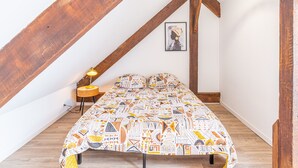 A bedroom with a sloped ceiling, a bed with patterned bedding, a bedside lamp, and a hanging light fixture. - Le Guindy - Localisation idéale (Treguier)