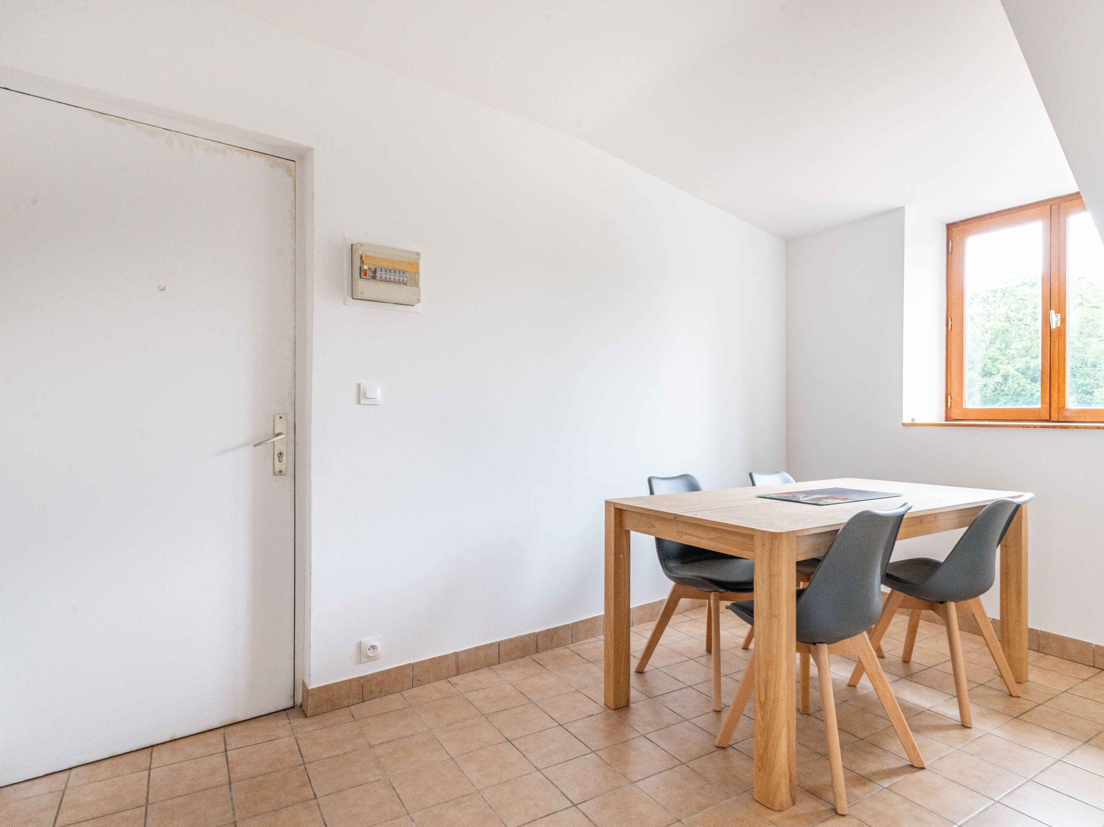 Un coin repas avec une table en bois et quatre chaises, une porte blanche, un interrupteur et une fenêtre donnant sur un paysage verdoyant.