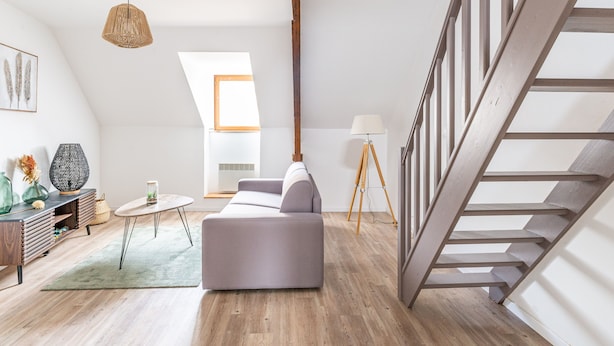 A spacious loft with a high ceiling, a wooden staircase, a grey sofa, a floor lamp, and a large window providing natural light. - Le Jaudy - Localisation idéale (Treguier)